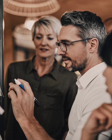Ein Mann mit Brille schreibt an einer Tafel, während zwei Frauen ihm aufmerksam zuschauen. Die Szene spielt sich in einem modernen Büro oder Besprechungsraum ab.