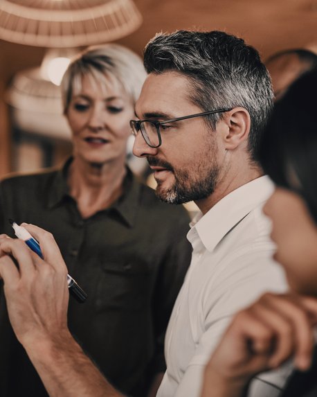 Ein Mann mit Brille schreibt an einer Tafel, während zwei Frauen ihm aufmerksam zuschauen. Die Szene spielt sich in einem modernen Büro oder Besprechungsraum ab.