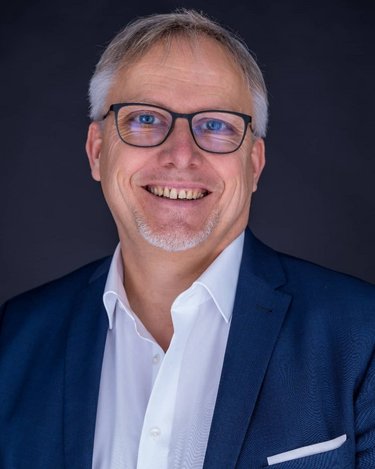 Ein lächelnder Mann mit Brille trägt einen blauen Anzug und ein weißes Hemd. Der Hintergrund ist dunkel und neutral.