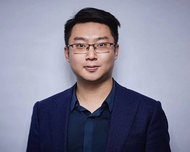 A man with glasses is wearing a dark suit and looking directly at the camera against a neutral background.