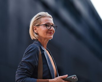 Eine blonde Frau mit Brille steht draußen und hält ein Smartphone in der Hand, während sie in die Ferne blickt.