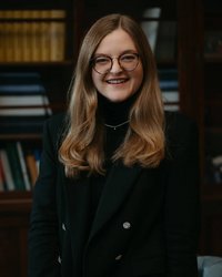 A smiling woman with long, curly hair and glasses is wearing a black outfit. Bookshelves are visible in the background.