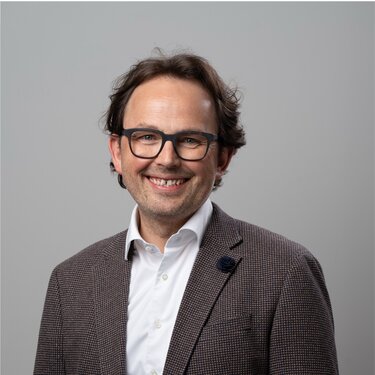 Mann mit Brille und lockigem Haar, trägt einen braunen Blazer und ein weißes Hemd, lächelt vor einem neutralen Hintergrund.