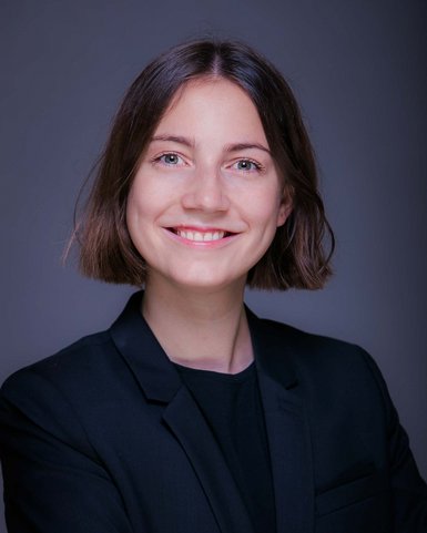 A smiling woman with shoulder-length brown hair is wearing a black blazer and stands with her arms crossed in front of a neutral background.