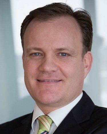 Ein Mann mit kurzen, braunen Haaren und einem Anzug lächelt in die Kamera. Er trägt ein gestreiftes Hemd und eine Krawatte.