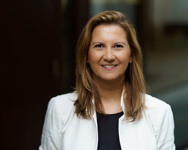 Eine lächelnde Frau mit langen, blonden Haaren trägt eine weiße Jacke und steht vor einem unscharfen Hintergrund.