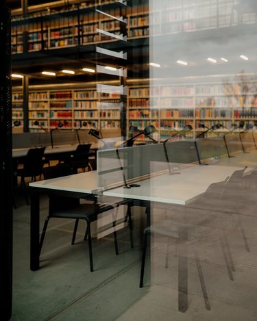 A modern reading room with tables and chairs, illuminated by desk lamps, with shelves full of books in the background.