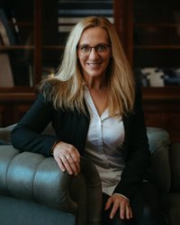 Eine blonde Frau mit Brille sitzt auf einem Sofa, trägt einen schwarzen Blazer und ein weißes Hemd, und lächelt freundlich.