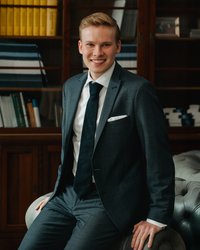 A young man in a gray suit is sitting smiling in an armchair in front of a bookshelf.