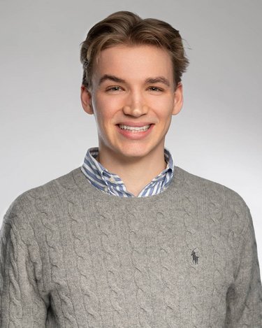 Ein junger Mann mit blonden Haaren lächelt, trägt einen grauen Pullover und ein gestreiftes Hemd darunter.