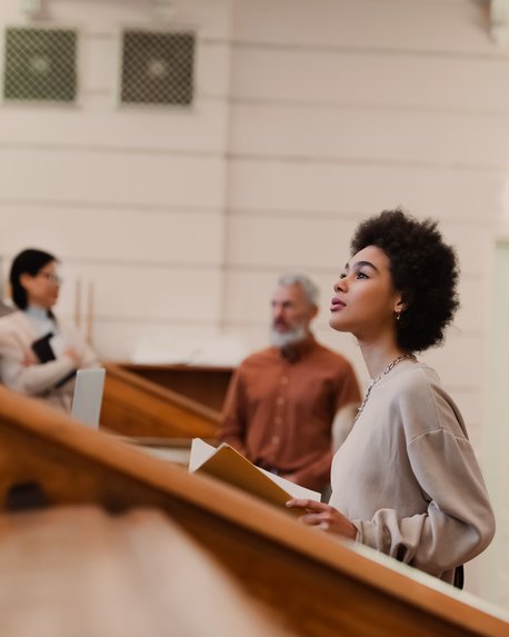 Eine Frau mit lockigem Haar steht an einer Treppe und hält Unterlagen in der Hand, während andere Personen im Hintergrund stehen.