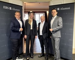 Vier Personen stehen vor zwei Banner mit der Aufschrift "EBS Alumni". Sie tragen formelle Kleidung und posieren freundlich.