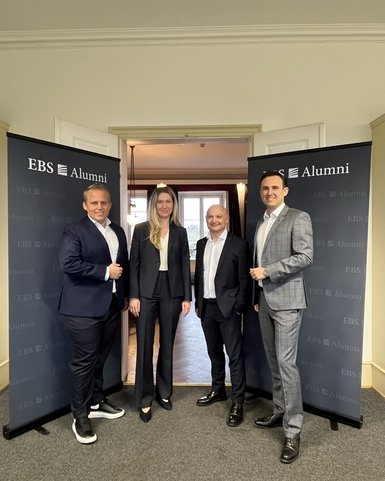 Four people stand in front of two banners that read 'EBS Alumni.' They are dressed in formal attire and are posing cheerfully.