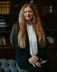 Eine Person mit langen, lockigen Haaren in einem schwarzen Anzug sitzt auf einem gepolsterten Sofa vor einem Bücherregal.