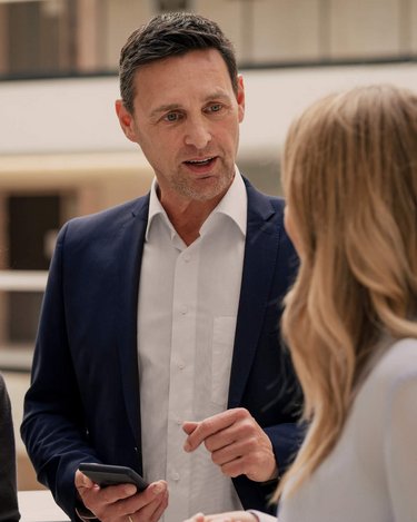 Drei Personen in formeller Kleidung stehen in einem modernen Innenraum. Eine Frau mit lockigem Haar hält ein Tablet, während ein Mann mit kurzem Haar und Anzug spricht. Eine weitere Frau hört aufmerksam zu.