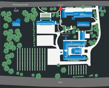 Site plan of an area with various buildings, including the main building, forum, and reception. The entrance area is marked. Surrounded by green spaces and vineyards, with street names indicated along the edge.