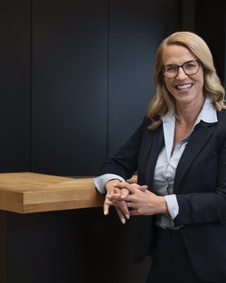 A woman with blonde hair and glasses is wearing a dark blue blazer over a light blue shirt. She stands in a modern, bright interior with glass walls and smiles warmly.