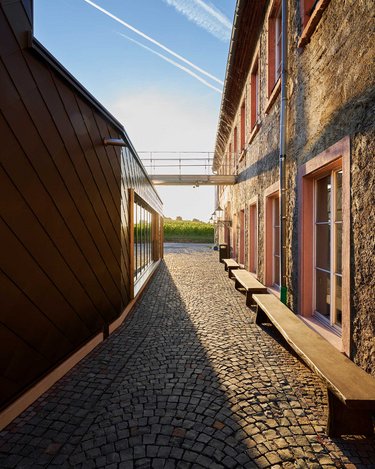 Ein schmaler, gepflasterter Durchgang zwischen zwei Gebäuden, eines mit einer modernen, dunklen Fassade und das andere mit einer traditionellen, hellen Steinmauer. Im Hintergrund ist eine Brücke sichtbar, die die beiden Gebäude verbindet.