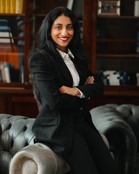 A woman with long black hair is wearing a black suit and smiling as she sits on a leather sofa with her arms crossed.
