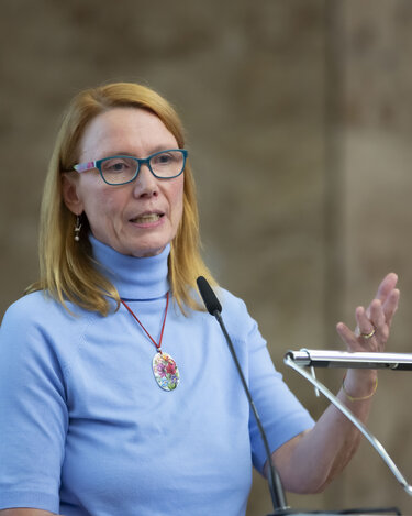Eine Frau mit blonden Haaren und Brille spricht am Rednerpult und gestikuliert. Sie trägt ein hellblaues Oberteil und eine Halskette.