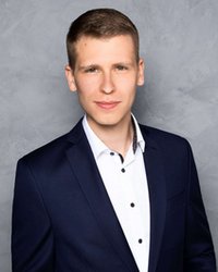 Ein junger Mann in einem dunklen Anzug mit weißem Hemd, der vor einem grauen Hintergrund steht und direkt in die Kamera schaut.