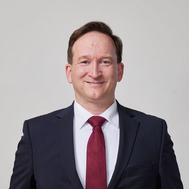 Man in a suit with a red tie, smiling and with his hands in his pockets, against a neutral background.