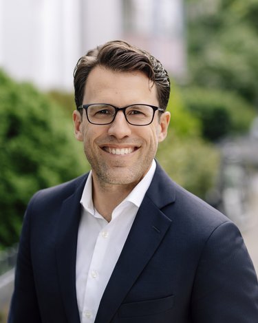 Mann mit Brille und Anzug steht lächelnd auf einem Gehweg, umgeben von grünen Sträuchern und einem unscharfen Hintergrund.