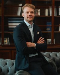 A man in a suit sits comfortably on a Chesterfield sofa, with his arms crossed and a friendly smile.