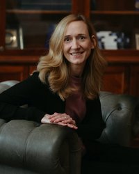 Eine Frau mit langen, lockigen Haaren sitzt auf einem Sessel und lächelt. Sie trägt einen schwarzen Pullover und eine lila Bluse.