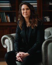 A person in a black suit is sitting in a green armchair, looking directly at the camera. Bookshelves are visible in the background.