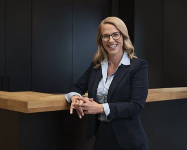 A smiling woman wearing glasses in a black suit stands at a wooden desk in a modern office.