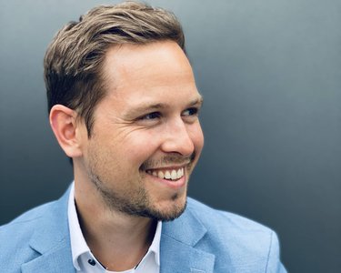 Ein Mann mit kurzen, braunen Haaren trägt ein hellblaues Sakko und ein weißes Hemd mit Knöpfen. Er lächelt und schaut leicht zur Seite, vor einem grauen Hintergrund.