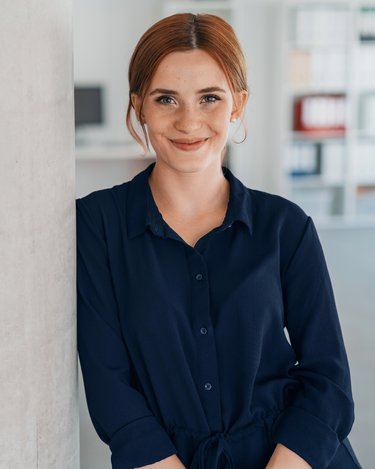 Junge Frau mit roten Haaren in einem blauen Hemd, die lächelnd an einer Betonwand in einem modernen Büro steht.