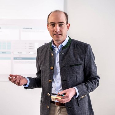 Ein Mann in traditioneller Kleidung steht vor einem Präsentationsboard und hält Notizen in der Hand, während er spricht.