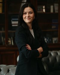 Eine Frau mit langen, dunklen Haaren steht lächelnd mit verschränkten Armen in einem eleganten, dunklen Blazer. Im Hintergrund sind Bücherregale und eine Couch sichtbar.