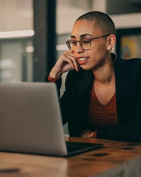 Eine Frau mit kurzen, rasierten Haaren und Brille sitzt an einem Tisch und schaut auf einen Laptop. Sie trägt ein schwarzes Blazer und ein rotes Oberteil, während sie nachdenklich mit der Hand an ihrem Kinn ruht.