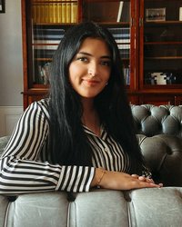 Eine Frau mit langen, schwarzen Haaren trägt eine gestreifte Bluse und sitzt entspannt auf einem gepolsterten Sofa.