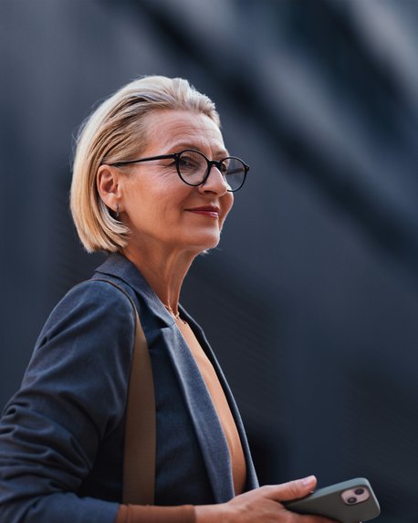 Eine blonde Frau mit Brille hält ein Smartphone in der Hand und schaut nachdenklich in die Ferne, gekleidet in einem blauen Blazer.