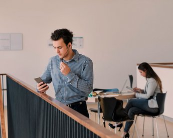 Student im Vordergrund am Smartphone; Studentin im Hintergrund am Tisch 