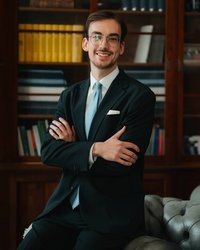 A young man in a suit and tie smiles as he sits on a sofa with his arms crossed. Bookshelves are visible in the background.