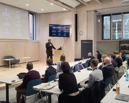 Ein Referent steht an einem Podium und spricht vor einem Publikum in einem modernen Veranstaltungsraum mit großen Bildschirmen.