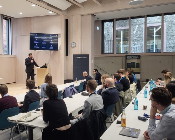 Ein Referent steht an einem Podium und spricht vor einem Publikum in einem modernen Veranstaltungsraum mit großen Bildschirmen.