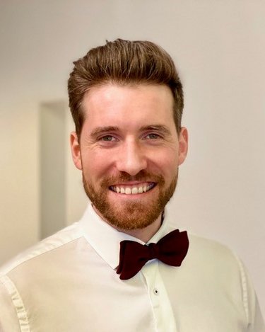 Ein Mann mit einem Bart und einer Fliege lächelt in einem eleganten Hemd. Der Hintergrund ist neutral und unscharf.