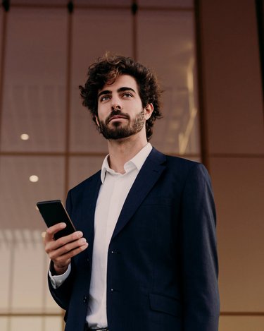 Ein Mann mit lockigem Haar trägt einen Anzug und hält ein Smartphone in der Hand. Er steht vor einem modernen Gebäude mit großen Fenstern.