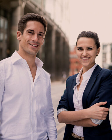 Zwei junge Erwachsene stehen in einer urbanen Umgebung. Der Mann trägt ein weißes Hemd, die Frau einen blauen Blazer. Beide lächeln und wirken selbstbewusst, während sie in die Kamera schauen.
