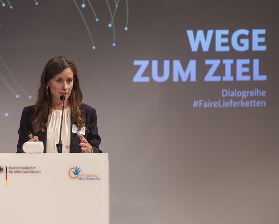 A woman speaks at the podium during an event titled 'Paths to the Goal' as part of the dialogue series #FairSupplyChains.