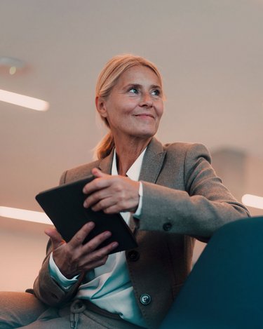 Eine Frau in einem grauen Anzug sitzt auf einem Stuhl und hält ein Tablet in der Hand. Sie blickt nachdenklich und hat lange, blonde Haare. Der Hintergrund ist modern und minimalistisch gestaltet.
