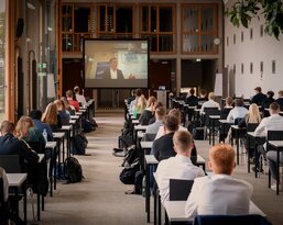 Eine Gruppe von Studierenden sitzt in einem Klassenzimmer, während sie einem Vortrag auf einem großen Bildschirm folgen. Die Anordnung der Tische ist in Reihen, und die Teilnehmer sind auf das Geschehen auf dem Bildschirm konzentriert.