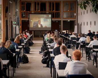 Eine Gruppe von Studierenden sitzt in einem Klassenzimmer, während sie einem Vortrag auf einem großen Bildschirm folgen. Die Anordnung der Tische ist in Reihen, und die Teilnehmer sind auf das Geschehen auf dem Bildschirm konzentriert.