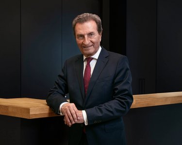 A man in a suit stands at a wooden bar, smiling, with a modern backdrop in dark colors.